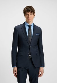 Marineblauer maßgeschneiderter Anzug mit texturiertem Finish, ergänzt durch ein hellblaues Hemd und eine gemusterte schwarze Krawatte. Das Einstecktuch hat blaue Akzente.