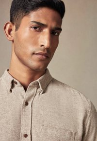 Beige skjorte med knaplukning lavet af tekstureret stof, spidse krave og en brystlomme. Brune knapper tilføjer detalje til designet.