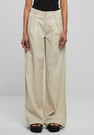 Beige Hosen mit weitem Bein, vorderen Falten, hoher Taille und Knopfverschluss, getragen mit schwarzen Plateau-Sandalen und einem schwarzen Top.