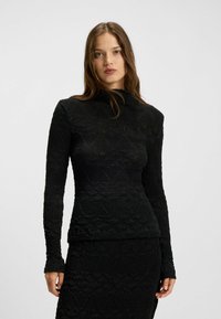 Schwarzes Langarmoberteil mit einem strukturierten Muster aus erhöhten Dreiecken und Wellen, mit hohem Kragen und taillierter Silhouette. Weicher Stoff.
