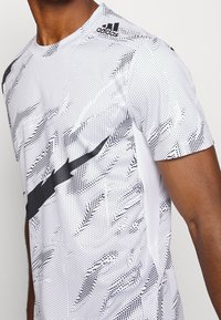 Camiseta atlética blanca con patrón de puntos negros y acentos de franjas diagonales negras. Presenta mangas cortas y un cuello redondo.