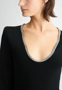 Schwarzes Kleidungsstück mit tiefem V-Ausschnitt und silbernem Strassbesatz am Halsausschnitt, glatte Textur und langen Ärmeln.