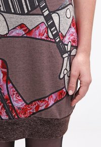 Vestido marrón de manga corta con un patrón floral de rosas rosas, contornos negros y un dobladillo texturizado marrón. Mano visible en el costado.