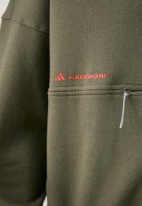 Olivengrøn sweatshirt med en glat tekstur, der har et rødt logo og lynlåslomme detaljer på ærmet.