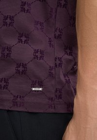 Dunkellila Hemd mit einem strukturierten Muster aus miteinander verbundenen Formen. Ein kleines Markenetikett ist am unteren Saum sichtbar.