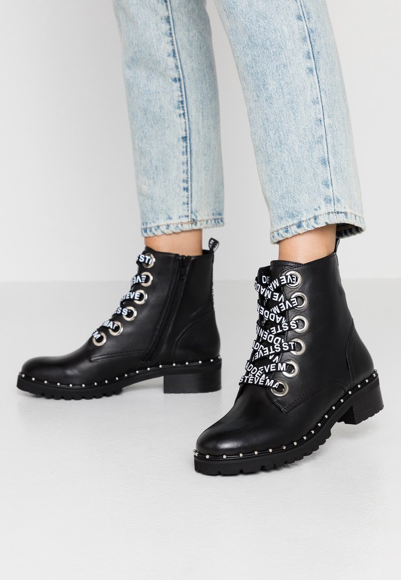 Bottines noires en cuir lisse avec des lacets décoratifs blancs, des œillets en métal argenté, une semelle cloutée et une fermeture éclair sur le côté.