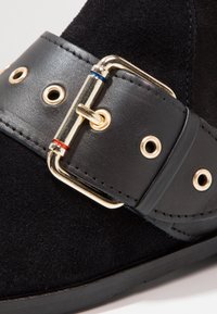 Detailní záběr na černou semišovou botu s širokým černým koženým popruhem a zlatým sponou, která má červené a modré smaltové akcenty.