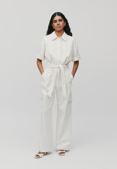 Hvid jumpsuit med korte ærmer, krave, bælte i taljen og brede ben. Har side lommer og diskrete manchetter. Båret med hvide sandaler.