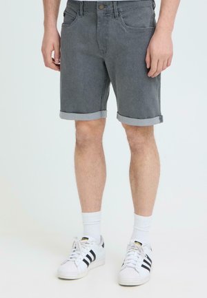 Homme portant un short en denim gris foncé retroussé, des chaussettes blanches montantes et des baskets Adidas blanches à bandes noires, debout devant un fond uni.