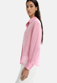 Jasnoróżowa koszula z długim rękawem, z kołnierzykiem na guziki, lekko przezroczystym materiałem i podwiniętymi mankietami. Posiada zaokrąglony dół i luźny krój.