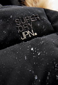 Tissu noir résistant à l'eau avec logo brodé "SUPER DRY JPN." et des gouttes d'eau sur la surface.