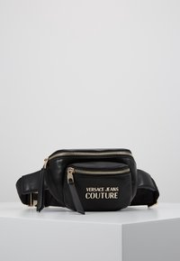 Černá kožená pásková taška se zlatými zipy a logem "Versace Jeans Couture" na přední kapse, vystavená na bílé ploše.