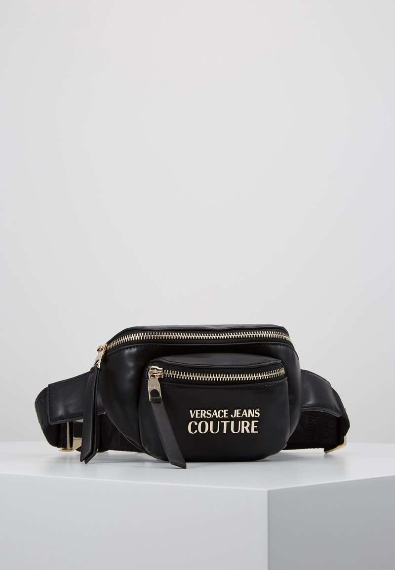 Černá kožená pásková taška se zlatými zipy a logem "Versace Jeans Couture" na přední kapse, vystavená na bílé ploše.