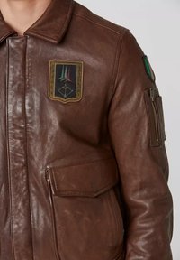 Primo piano di una giacca di pelle marrone con una toppa delle Frecce Tricolori sul petto e una piccola tasca con cerniera con una toppa della bandiera italiana sulla manica.