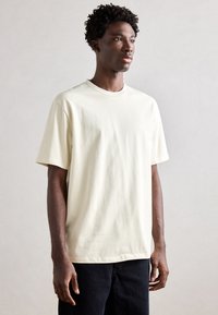 Tricou din bumbac de culoare crem, cu guler rotund, mâneci scurte și o croială lejeră, purtat cu blugi din denim închis. Textura materialului este simplă și netedă.