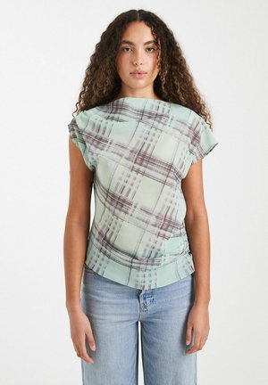 Kvinde med krøllet hår iført en lysegrøn top med korte ærmer med abstrakt ternet design og lyseblå jeans, stående mod en ensfarvet baggrund.