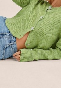 Hellgrüner, gerippter Cardigan mit weitem Ausschnitt und langen Ärmeln, versehen mit edelsteinartigen Knöpfen und einer weichen Strickstruktur, kombiniert mit hoch geschnittenen Jeans.