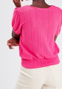 Blouse rose, texturée, avec des manches bouffantes, présentant une taille côtelée et un motif à œillets. Portée avec un pantalon clair taille haute.