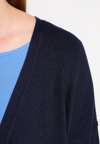 Námořnický kardigan s žebrovanou texturou a otevřeným předním dílem, přeložený přes světle modrou košili. Zaměřuje se na detaily výstřihu a rukávů.