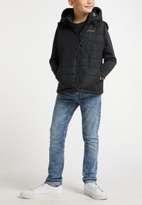 Gilet imbottito nero con zip frontale e cappuccio, indossato sopra una maglietta scura. Abbinato a jeans in denim chiaro e sneakers bianche.