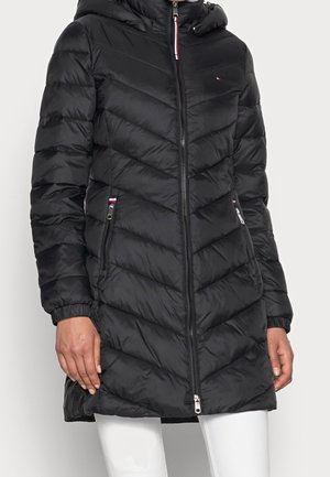 Veste longue matelassée noire avec capuche, fermeture éclair frontale, poches latérales zippées, et logo discret rouge, blanc et bleu sur la poitrine et les poches.