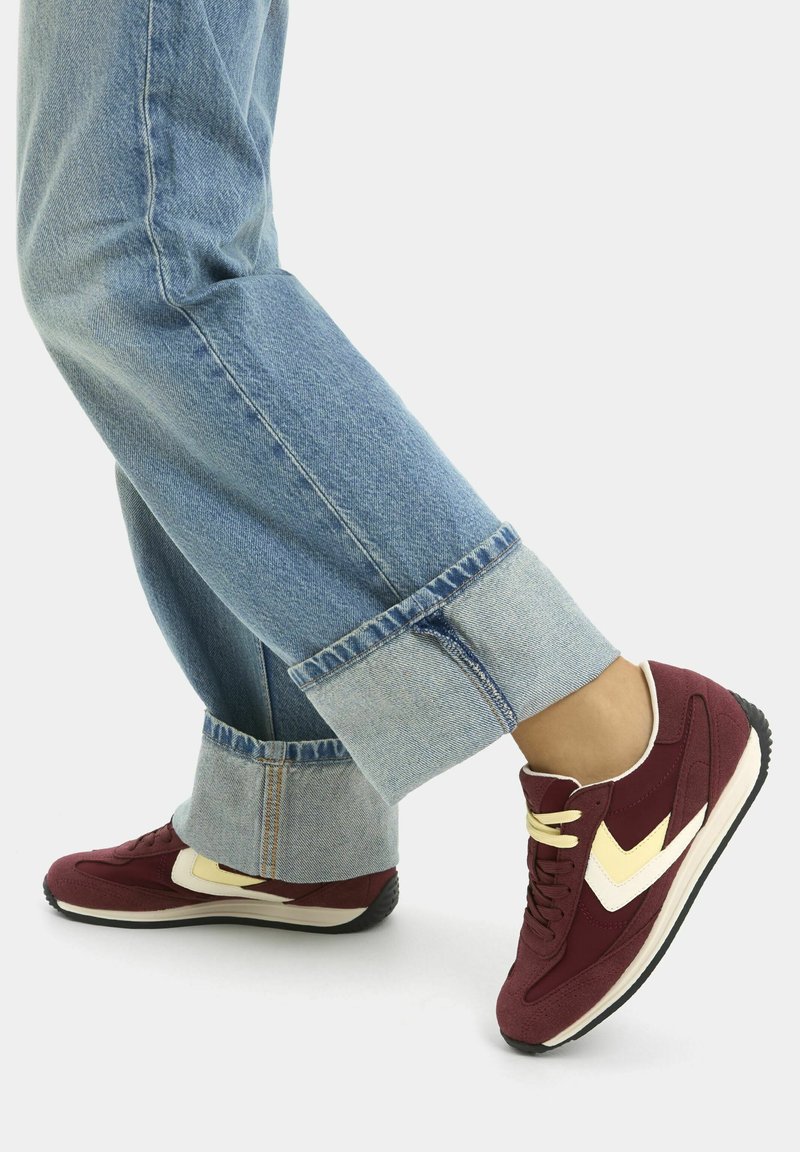 Personne portant un jean bleu clair retroussé et des baskets bordeaux avec des accents crème et blanc, debout sur un fond clair.