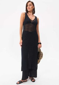 Schwarzes gestricktes Maxikleid mit V-Ausschnitt, das ein offenes Strickmuster aufweist. Kombiniert mit schwarzen weit geschnittenen Hosen und geflochtenen Rutsch-Sandalen.