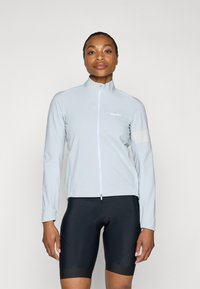 Light blue cycling jacket with a high collar, full-front zipper, and reflective accents. Worn over black cycling shorts. Smooth texture.