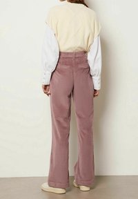 Ljust beige stickad väst över en vit långärmad skjorta, tillsammans med rosa kordjeans med vida ben och krämfärgade sneakers.