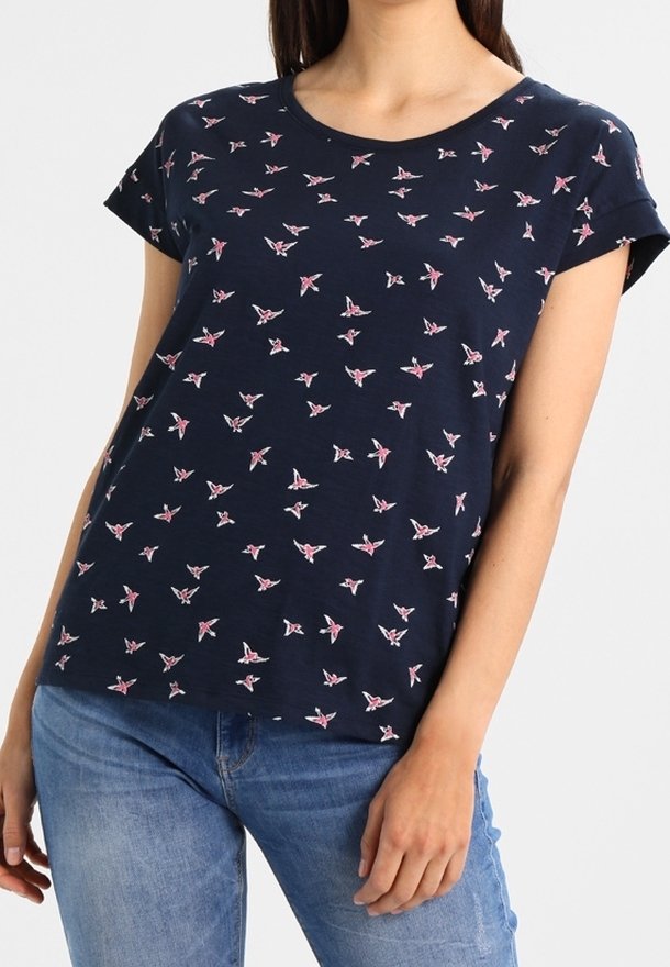 Kvinna iklädd en marinblå kortärmad t-shirt med små mönster av rosa och vita fåglar, tillsammans med blå jeans, stående mot en vit bakgrund.