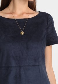Námorně modrý semišový top s krátkými rukávy, kulatým výstřihem a texturovaným povrchem. Je doplněn zlatým náhrdelníkem s kulatým přívěskem.