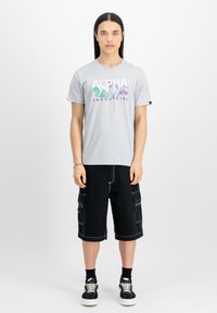 Šedé tričko s krátkým rukávem a fialovo-zeleným grafickým nápisem "ALPHA INDUSTRIES." Kombinováno s černými cargo šortkami a černými teniskami.
