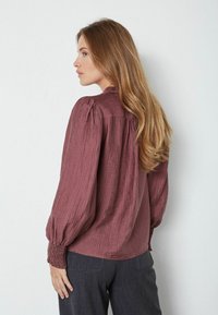 Bordó textúrájú blúz hosszú puffos ujjakkal és ráncolt mandzsettákkal, laza szabással és enyhén redőzött hátrészzel.