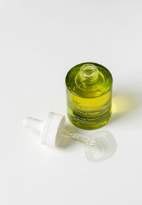 Grüne Glasflasche mit einem weißen Tropfverschluss, die ein klares Serum enthält. Das Etikett zeigt Text in mehreren Sprachen. Tropfen auf der Oberfläche.