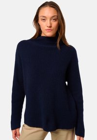 Pull à col roulé en tricot navy avec une texture côtelée, des manches longues et une coupe décontractée, associé à un pantalon de couleur claire.