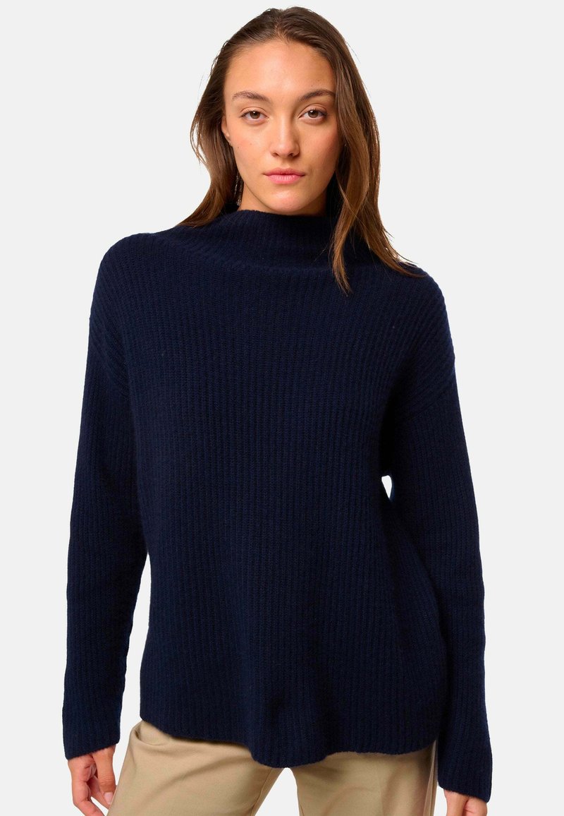 Pull à col roulé en tricot navy avec une texture côtelée, des manches longues et une coupe décontractée, associé à un pantalon de couleur claire.