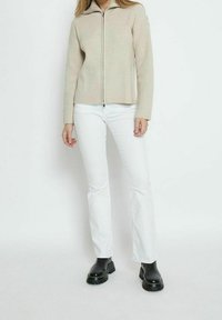 Beige ribbad zip-up tröja med hög krage, kombinerad med vita utsvängda jeans och svarta klumpiga klackboots. Enkel, modern design.