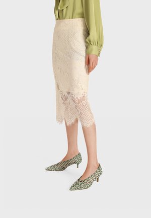 Donna che indossa una gonna midi beige in pizzo abbinata a scarpe verde con motivo, a punta e con tacco basso, e una blusa verde a maniche lunghe.