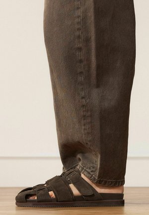 Fuß mit dunkelbraunem Wildleder-Sandalen mit offenem Zehenbereich und verstellbaren Riemen, kombiniert mit hochgekrempelter brauner Freizeithose auf hellem Boden.