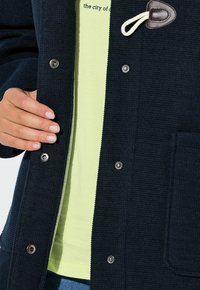 Tmavě modrý kabát s texturovaným povrchem, zapínáním na patentní knoflíky a uzavíráním na očko a knoflík. Má přední kapsu a je nošen přes světle žlutou košili s potiskem.