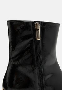 Gros plan d’une cheville de botte noire brillante avec une fermeture éclair verticale noire et un curseur doré sur fond clair.