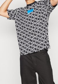 Svart t-shirt med vita, upprepande abstrakta mönster och en blå etikett. Kortärmad, rundhalsad design, tillverkad av lättviktigt material.