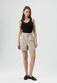 Zwart tanktop gecombineerd met beige shorts met hoge taille, voorzien van plooien en een omgeslagen zoom. Model draagt zwarte loafers met een gouden accent.