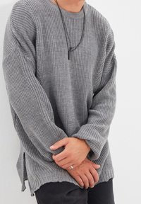 Pull gris en maille avec une coupe décontractée, manches longues, fentes latérales et motif texturé. Porté avec un pantalon noir.