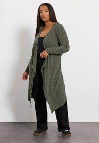 Oliwkowy długi kardigan z otwartym przodem, prążkowaną fakturą i korzystnie układającym się krojem. Połączony z czarną bluzką i czarnymi spodniami.