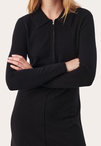 Robe noire à manches longues et au col classique. Présente une fermeture éclair sur le devant et une silhouette cintrée avec un tissu doux et texturé.