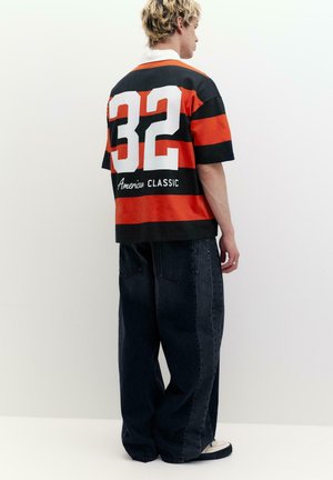 Osoba w czarno-czerwonej koszulce w paski z numerem 32 i napisem "American Classic", w połączeniu z luźnymi czarnymi jeansami i białymi tenisówkami.