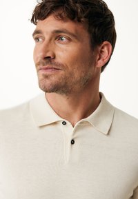 Beżowa koszulka polo z teksturowanej tkaniny, z listwą guzikową z dwoma czarnymi guzikami i klasycznym kołnierzykiem. Zbliżenie na dekolt.