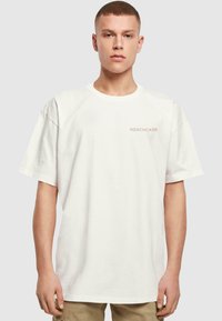 Hvid bomulds t-shirt med korte ærmer og rund halsudskæring. Har "MERCHC•DE" trykt i lys brun på brystet.
