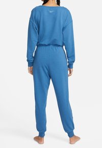Femme avec des cheveux tressés portant un sweat-shirt Nike bleu et un pantalon de jogging assorti, se tenant pieds nus tournée vers le dos sur un fond blanc.